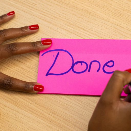 Close-up of a hand writing 'Done' on a pink sticky note with red nail polish.